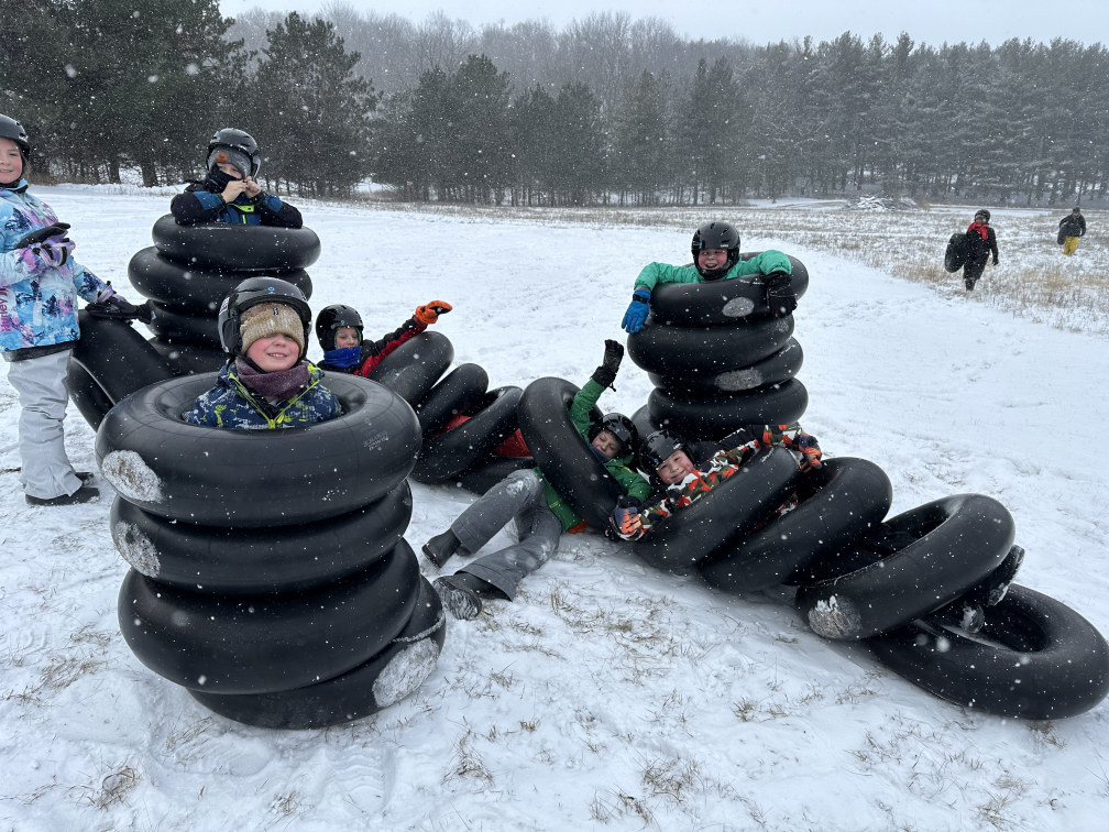 Several kids are standing in the snow in stacks of inflatable tubes for tubing, each smiling and wearing full winter gear.